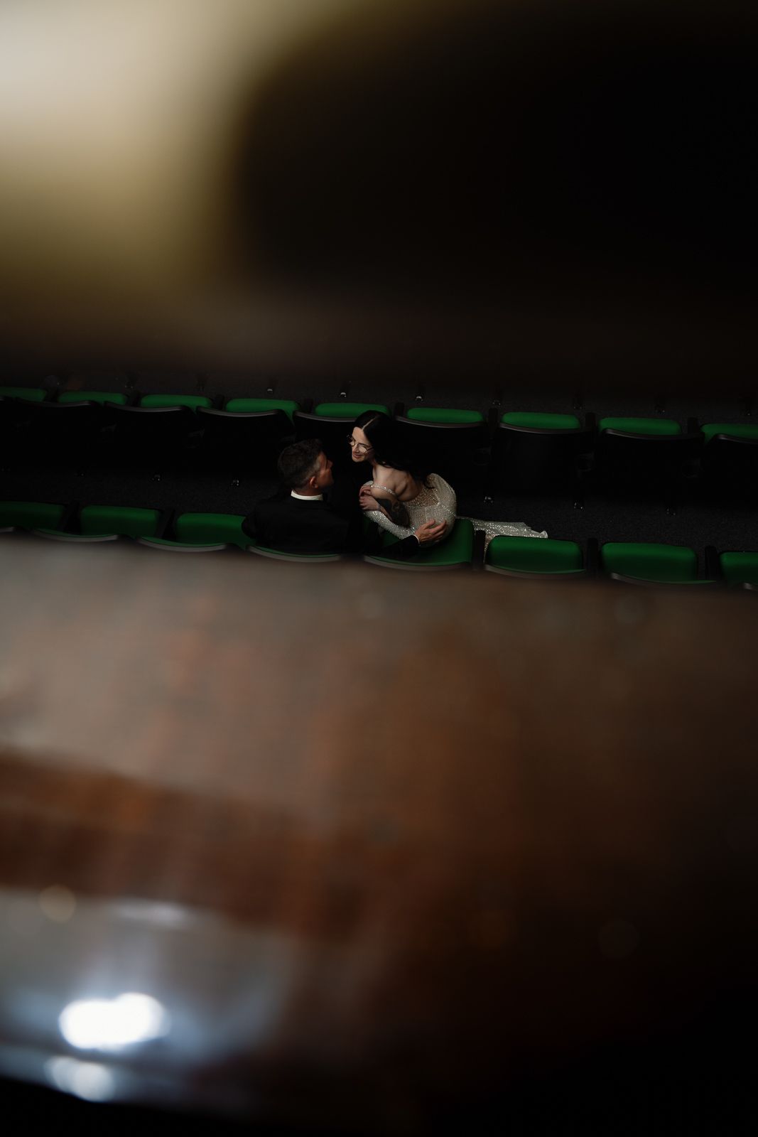 Couple sitting close together in dimly lit theater with green seats visible in the background.