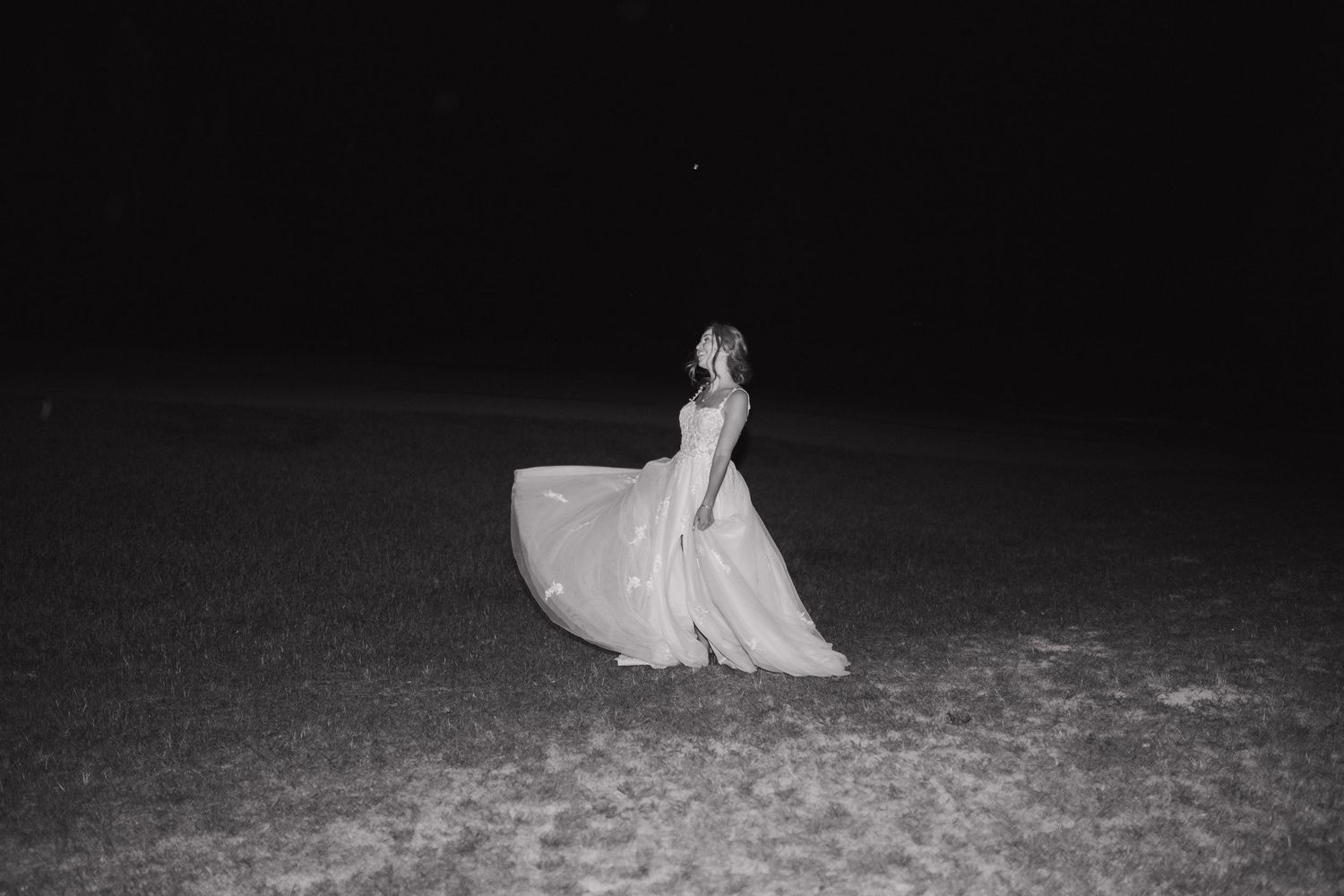 A figure in a flowing white dress twirls in darkness on a sandy beach, captured in black and white photography.