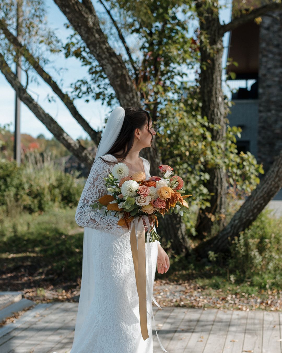 Bride holding autumn-colored bouquet poses on outdoor walkway surrounded by trees.