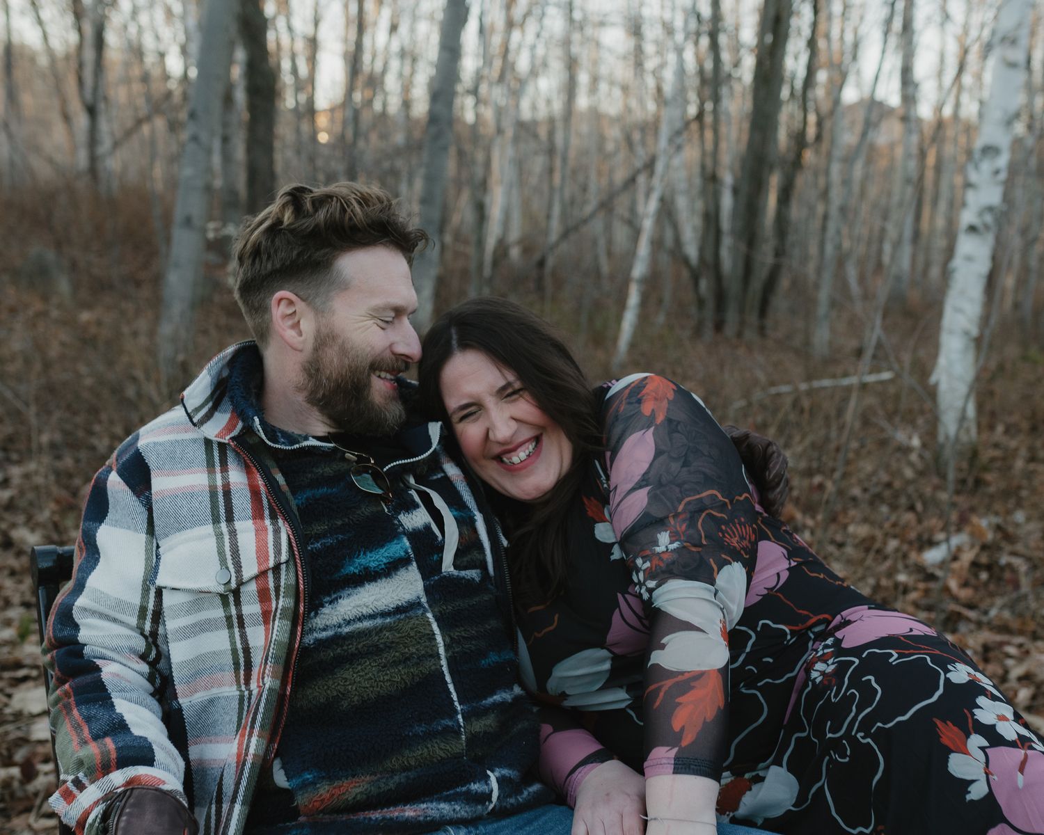 Couple in plaid shirts sharing tender moments while sitting among fallen autumn leaves in a wooded area.
