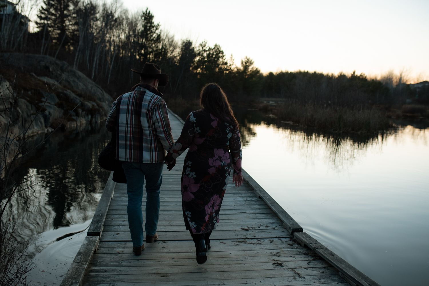 People walking on a wooden boardwalk path along a tranquil lake during sunset with trees in the background.