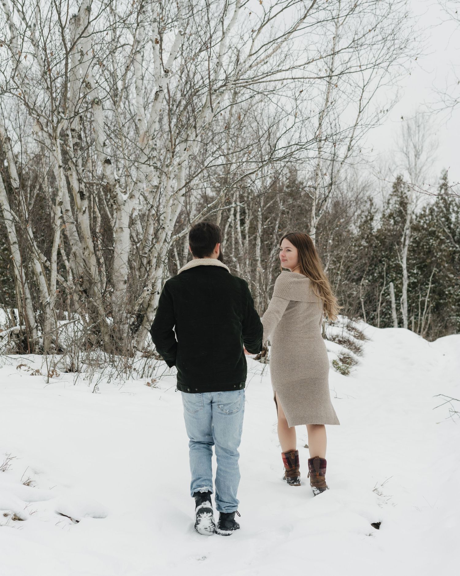 A couple walks through a snowy forest landscape holding hands.