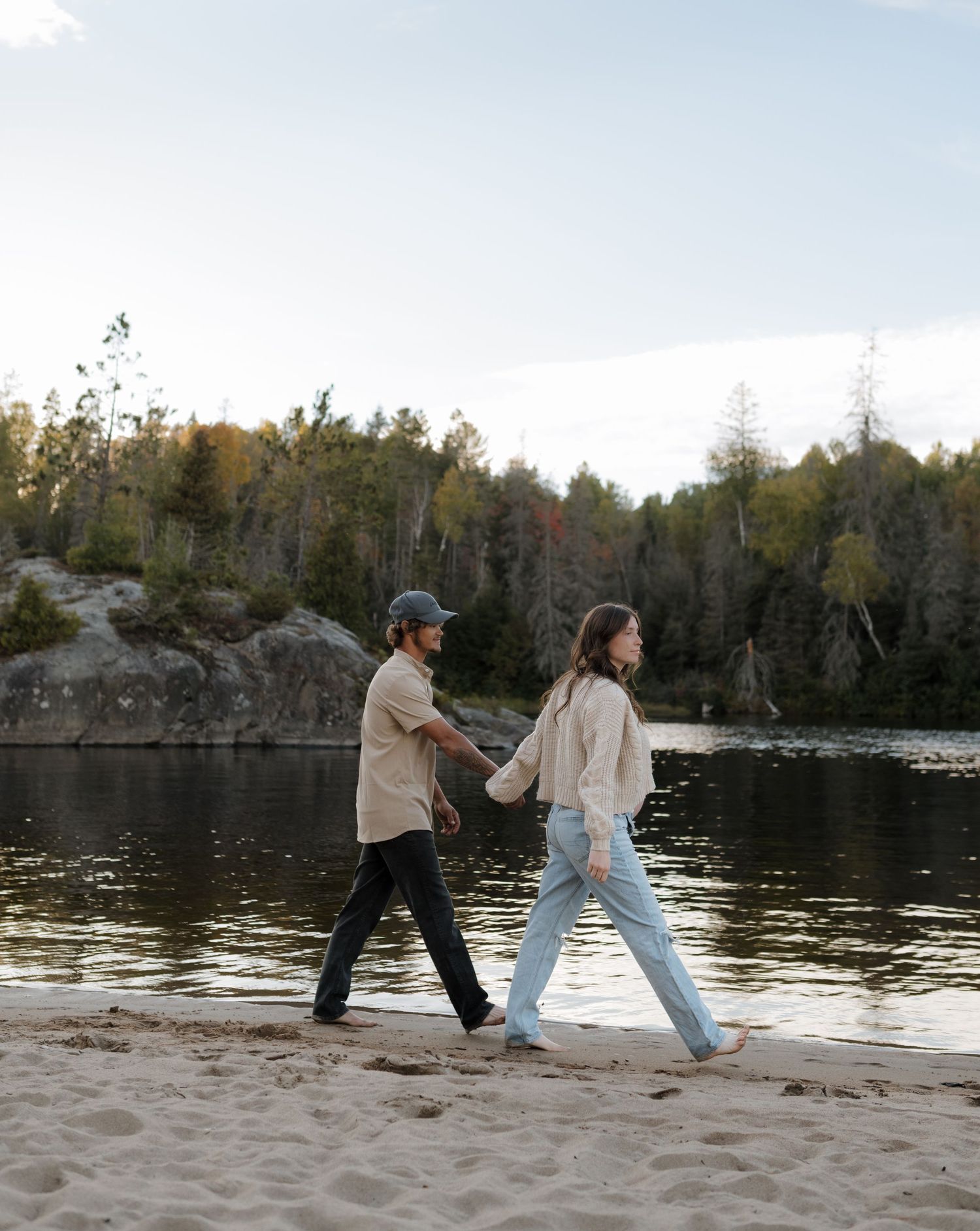 Two people dance playfully on a sandy beach with a lake and forest behind them.
