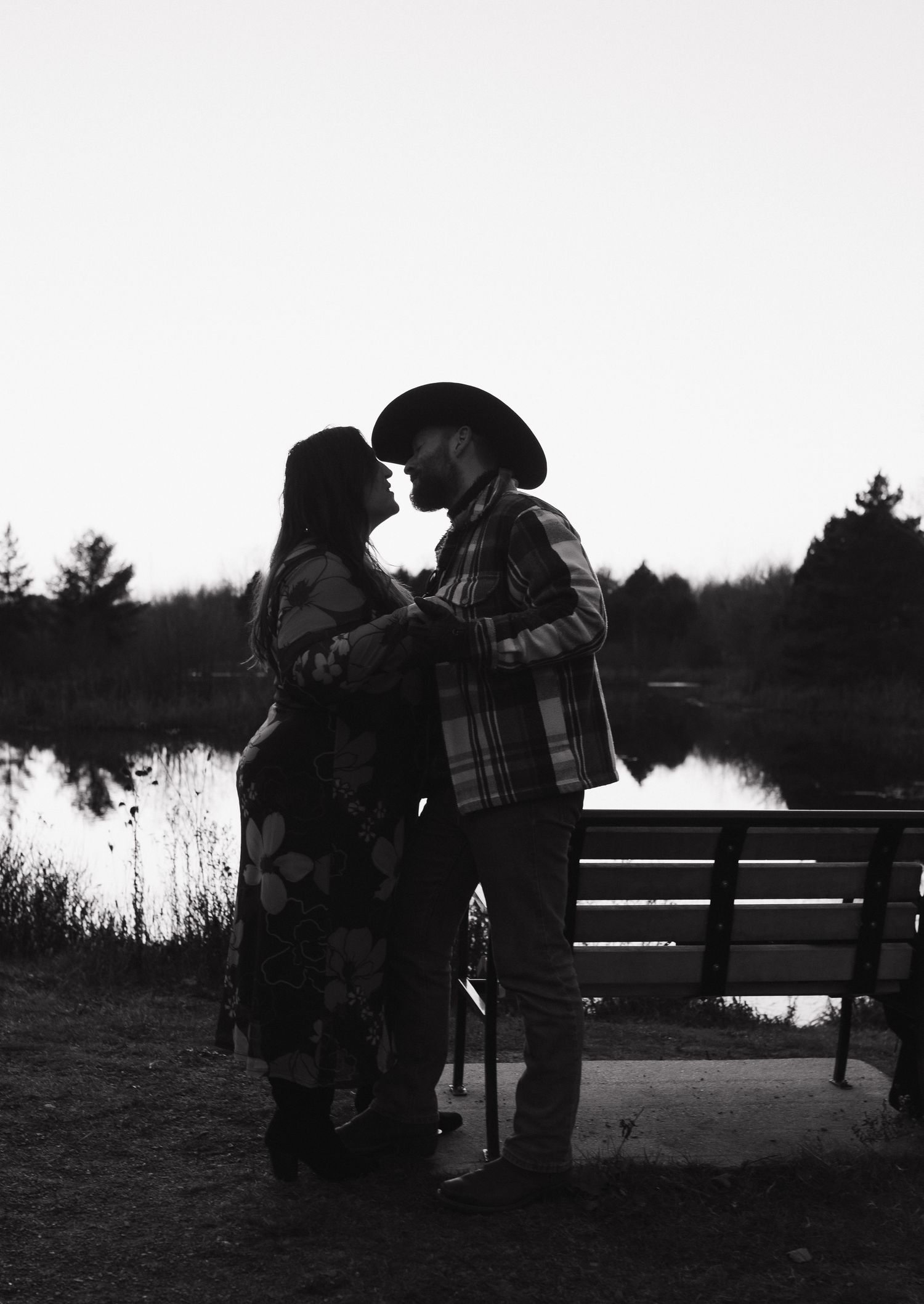 A silhouetted couple shares an intimate moment by a lake at dusk with a bench nearby.