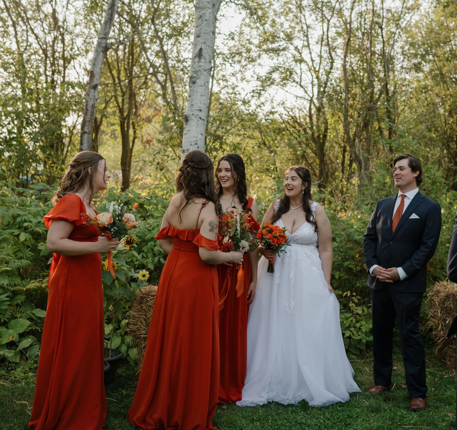 Bride in white gown with bridesmaids in red dresses holding bouquets outdoors at a fall wedding ceremony.