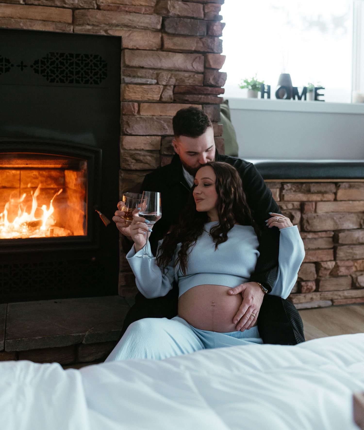 Pregnant couple embracing by a glowing stone fireplace, man kissing womans head.