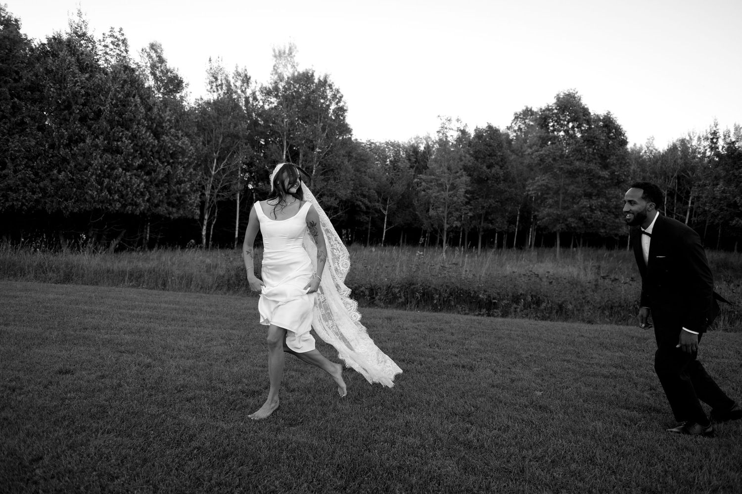 Black and white photo of a bride jumping joyfully in a field while groom watches, trees in background.