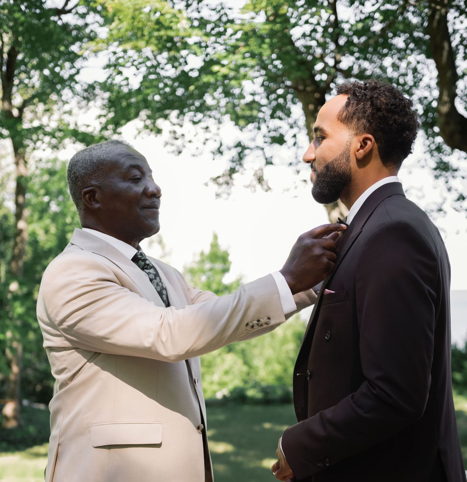 Older man in beige suit adjusts grooms bow tie outdoors before wedding ceremony.