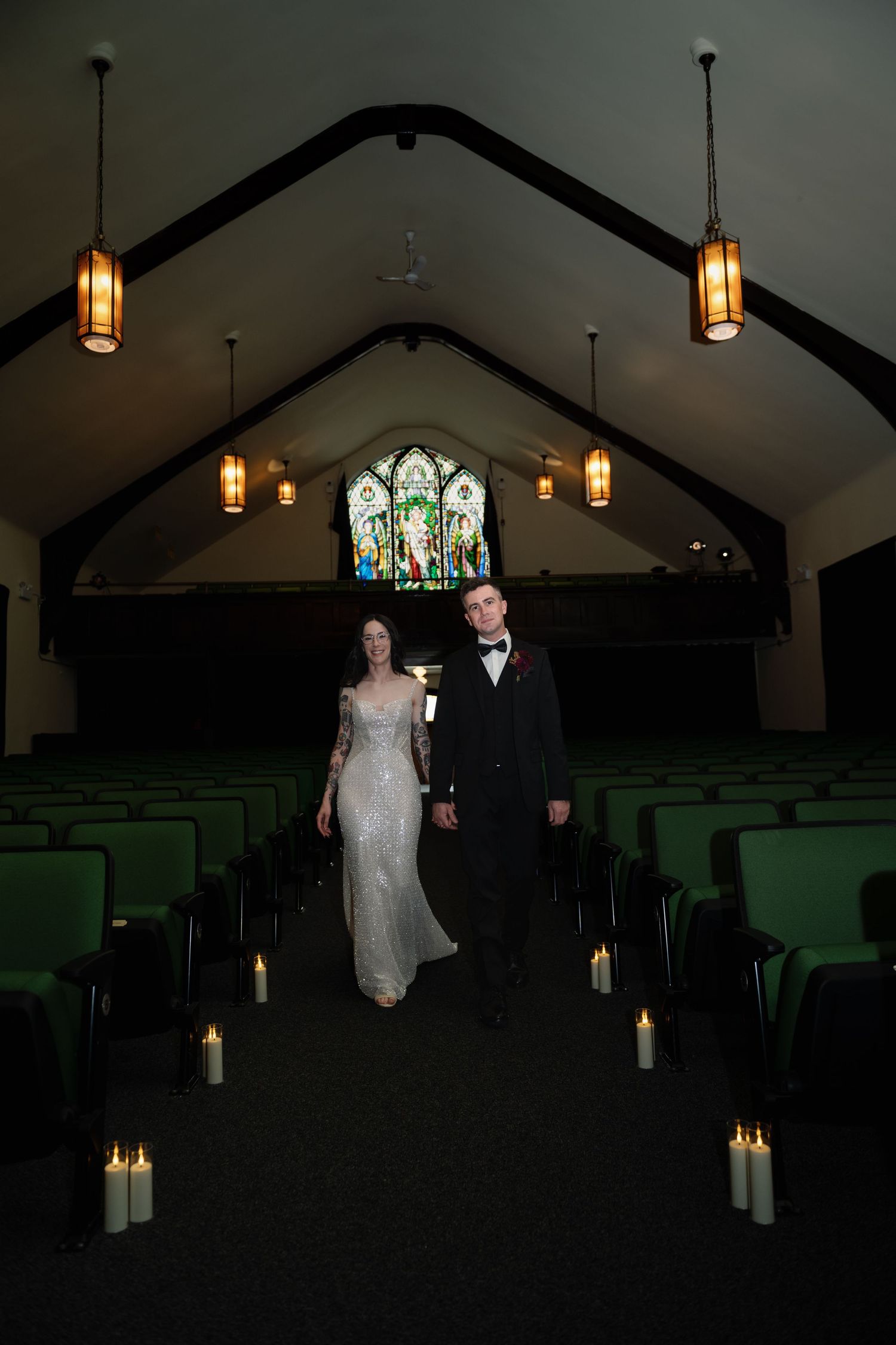 Bride and groom walk down candlelit church aisle with stained glass window backdrop in dimly lit chapel.