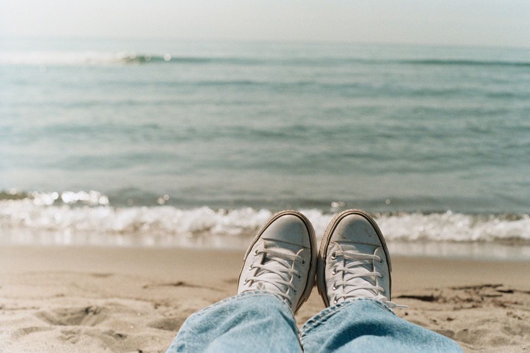 Person relaxing on sandy beach wearing blue jeans and gray sneakers with ocean waves in the background.