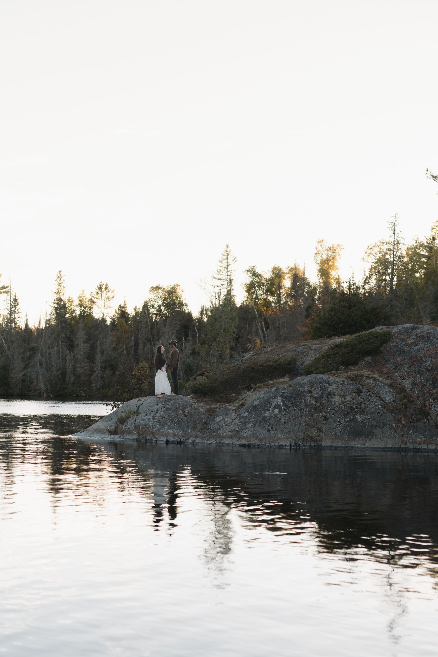 Scenic lakeside view at dusk with rocky cliffs and evergreen trees reflected in calm waters.