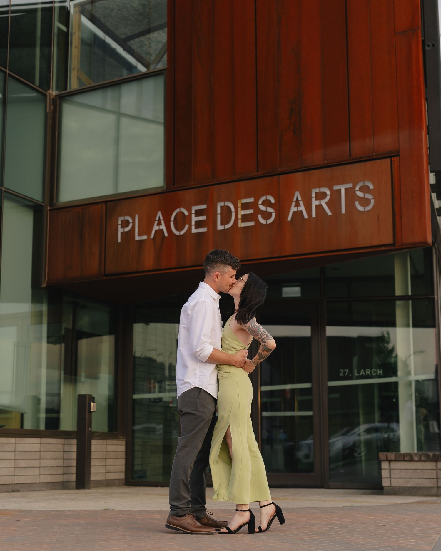 Couple sharing a romantic kiss in front of Place des Arts building, woman in green dress and man in white shirt.