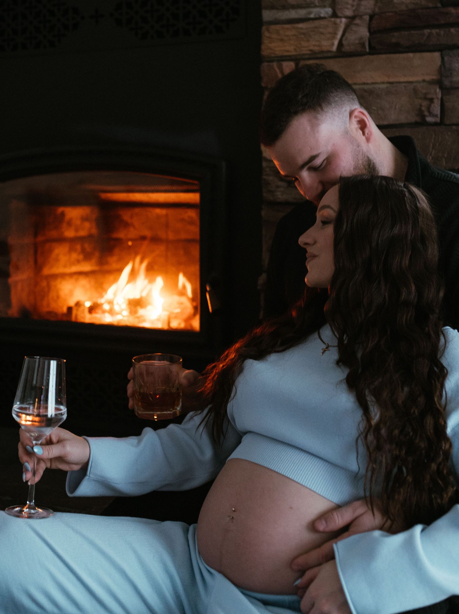 Pregnant couple embracing by a glowing fireplace, holding wine glasses in a cozy romantic setting.