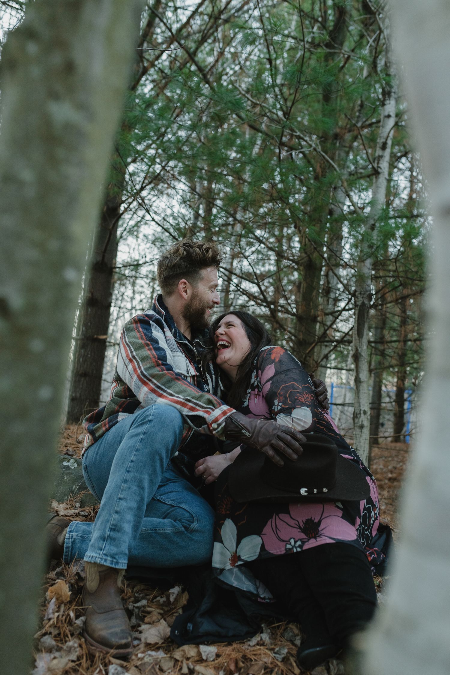 A couple shares a joyful moment while sitting on fallen leaves in a misty forest during autumn.
