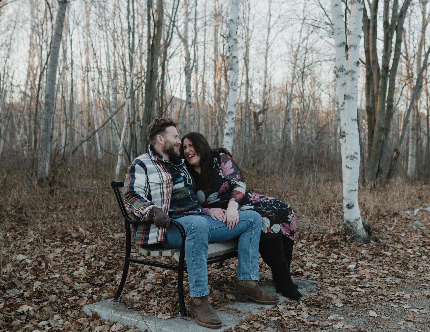 A romantic couple sits together on a rustic wooden bench surrounded by bare autumn trees in a forest setting.