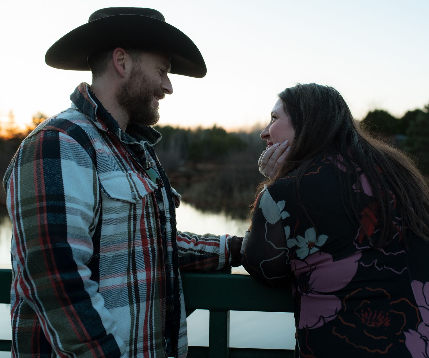 A romantic couple shares an intimate moment by a lake at sunset wearing casual western attire.