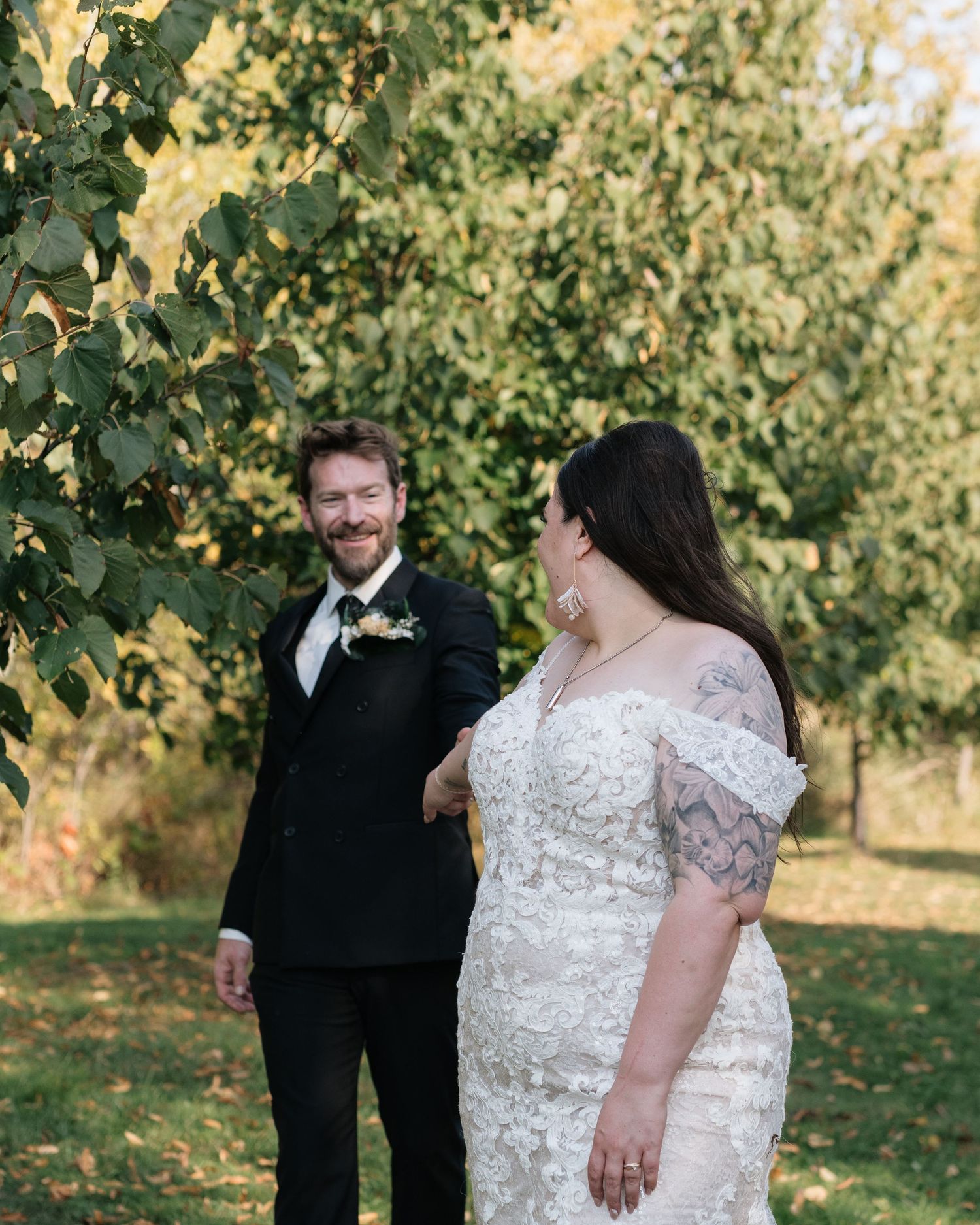 A bride in a lace gown and groom in a black tuxedo share a romantic first look moment outdoors among autumn trees.