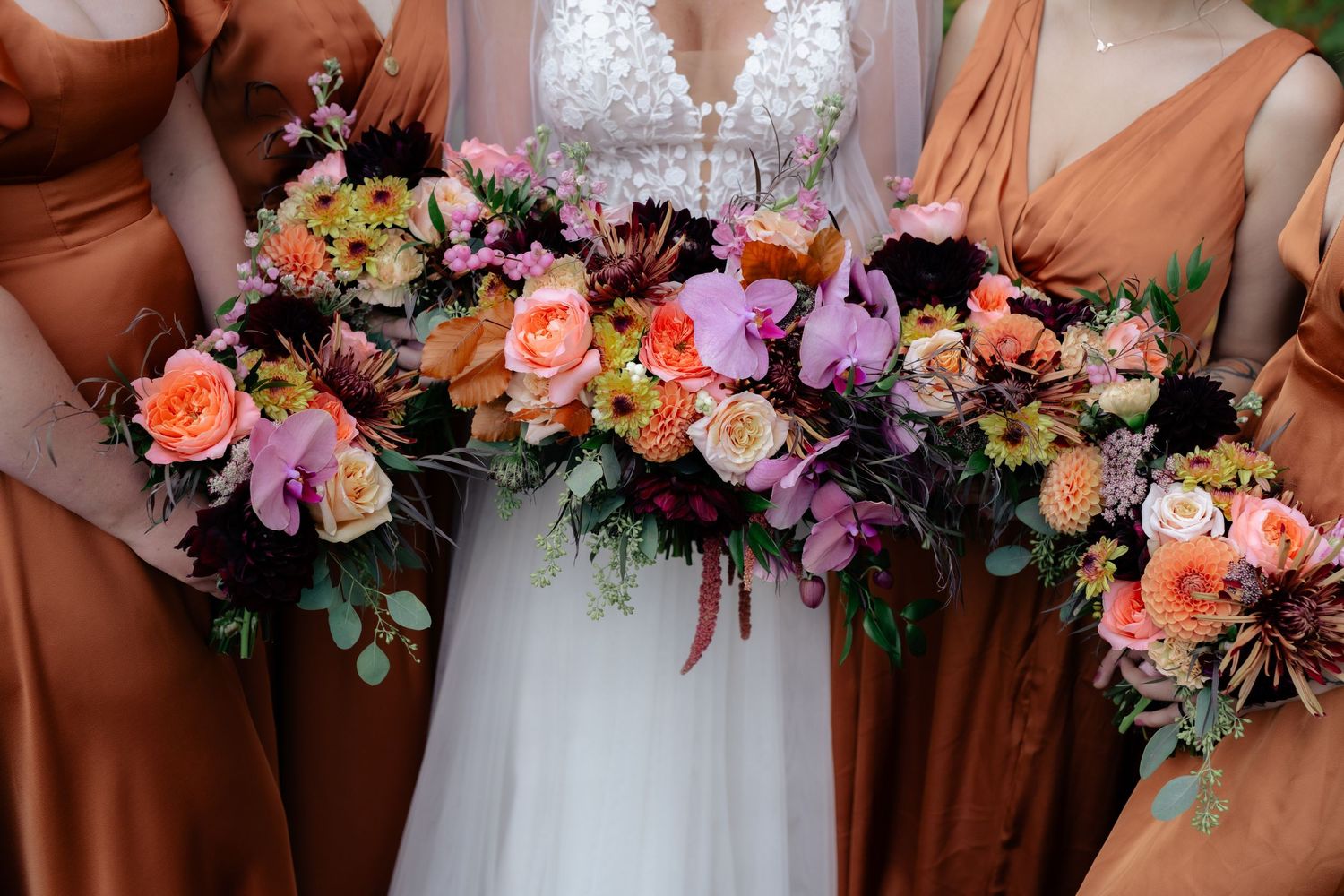 Bride and bridesmaids in copper dresses holding lush colorful wedding bouquets with roses, orchids, and greenery.