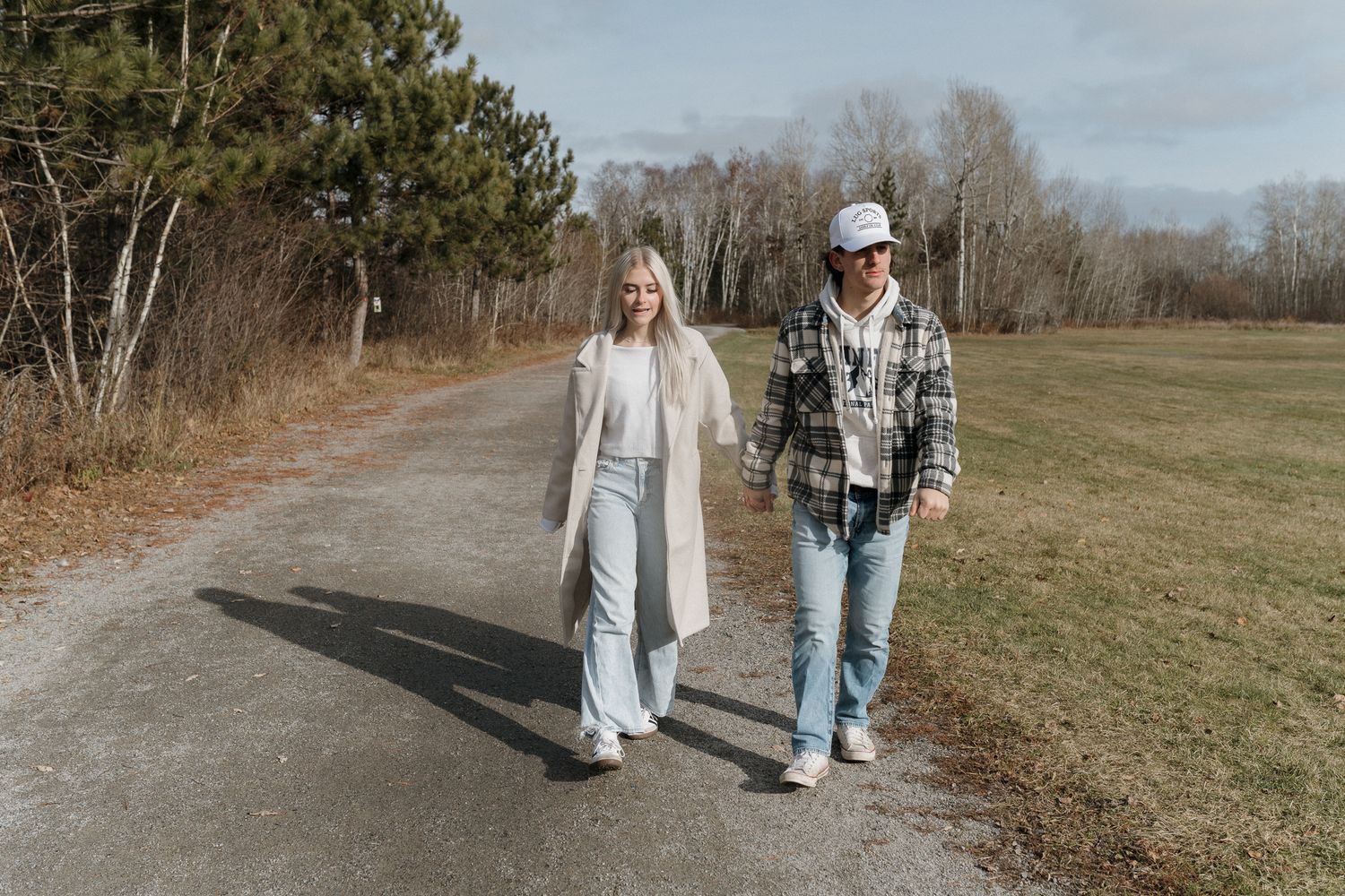 Couple holding hands and walking on a paved path through an open field on a sunny autumn day.