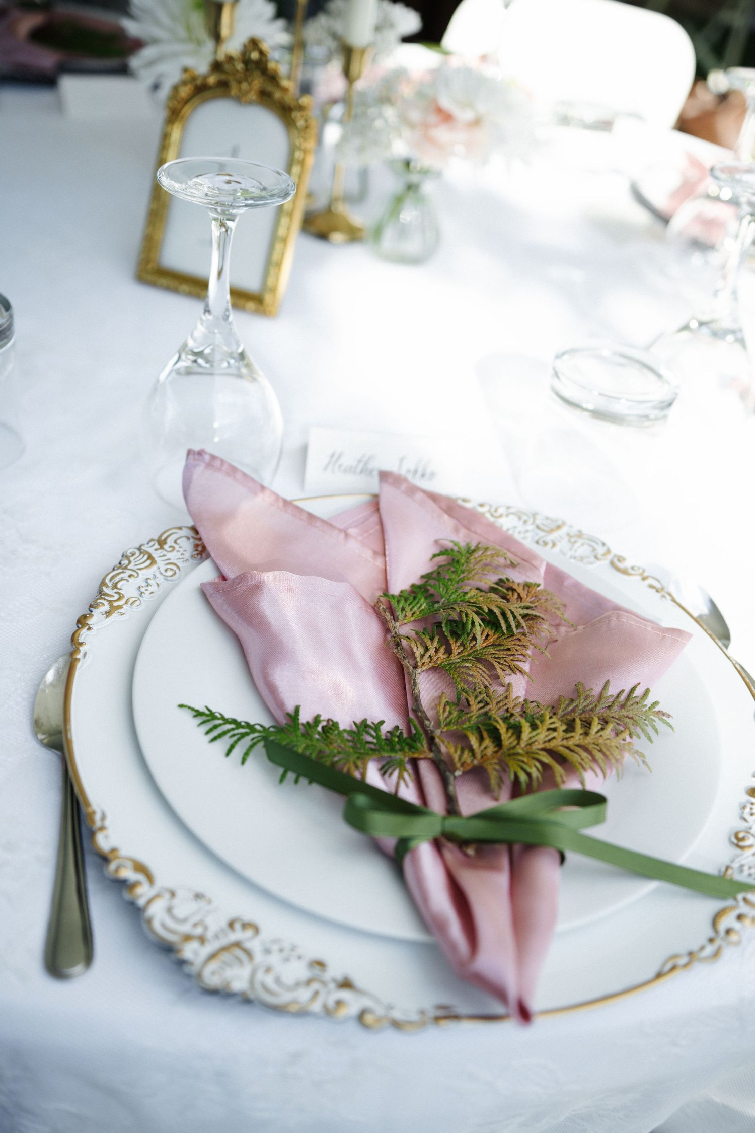 Elegant wedding table setting with pink silk napkin, greenery, gold-rimmed charger plate, and crystal glassware on white linen.