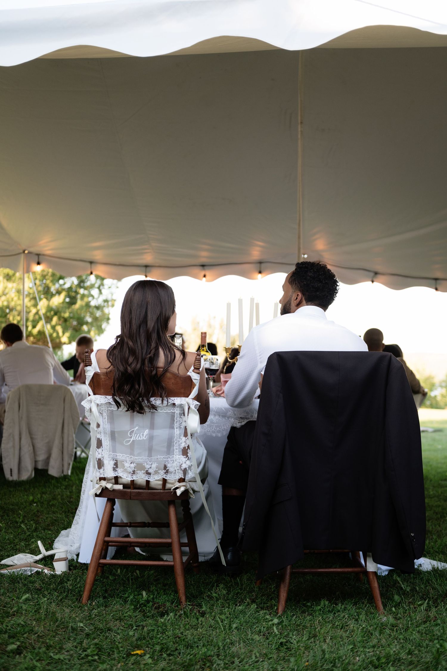 Bride and groom seated at outdoor wedding reception table under white tent, viewed from behind.