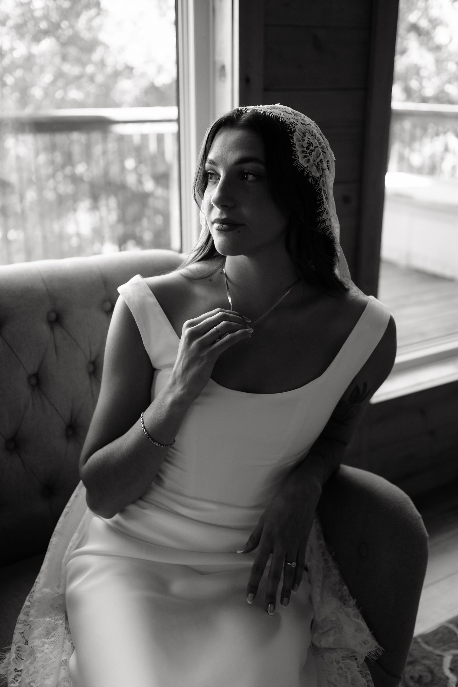 Black and white portrait of a bride in a white gown and lace headpiece seated by a window.