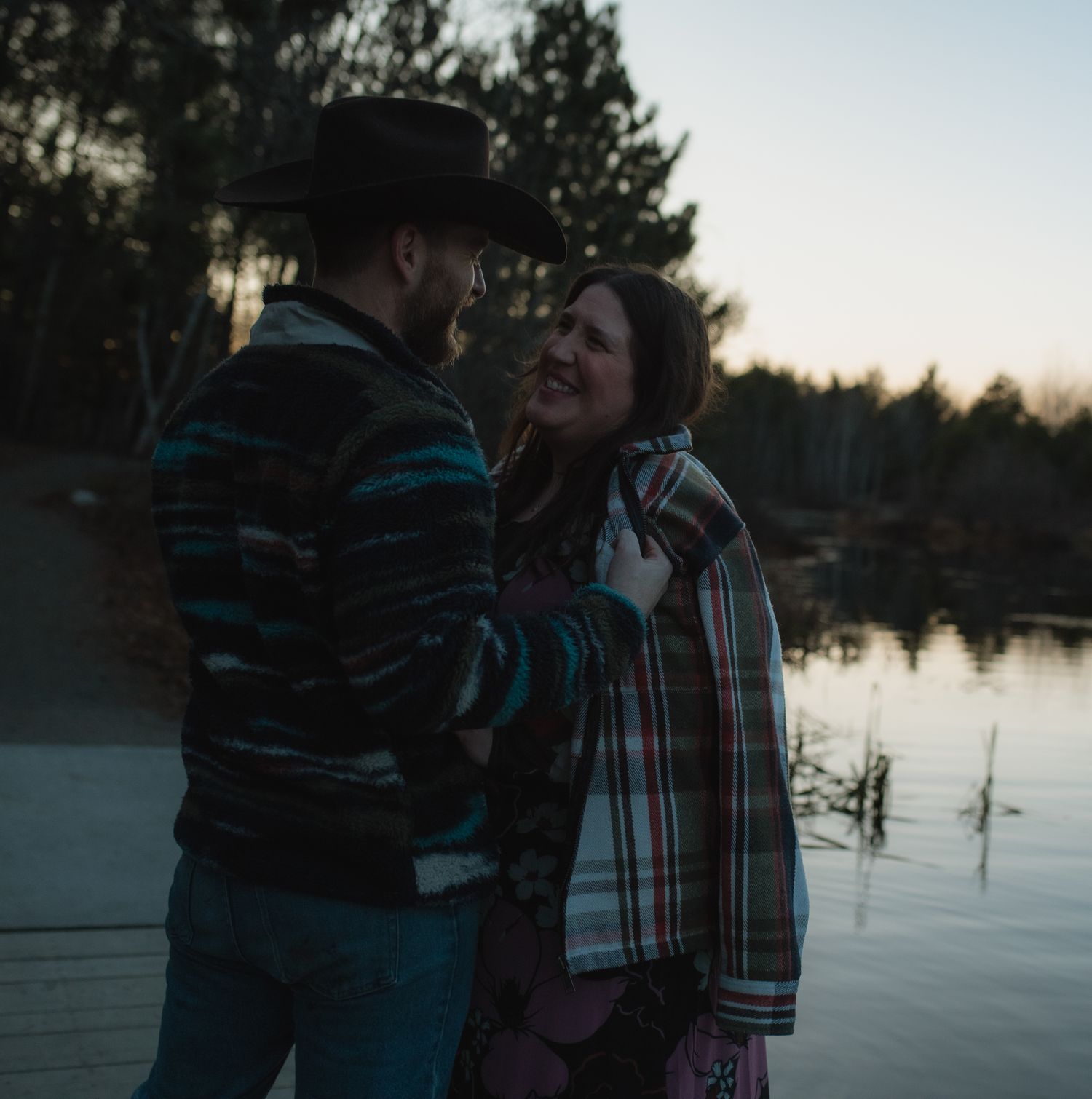 Couple embracing by a serene lake at sunset with trees silhouetted in the background.