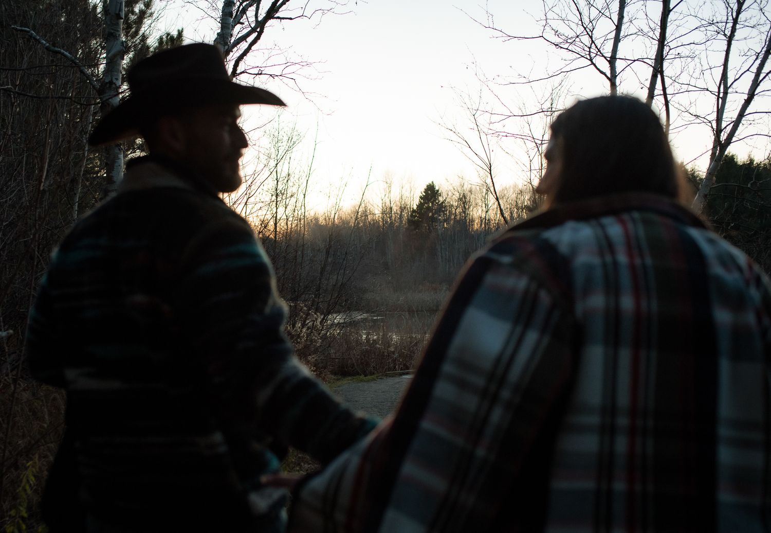 Silhouetted figures in plaid jackets stand against a dusky sky during sunset.