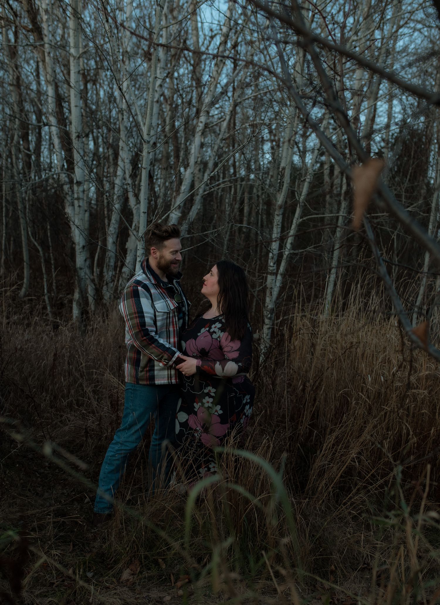 Two people embrace while standing in a wooded area with bare trees and brown grass.