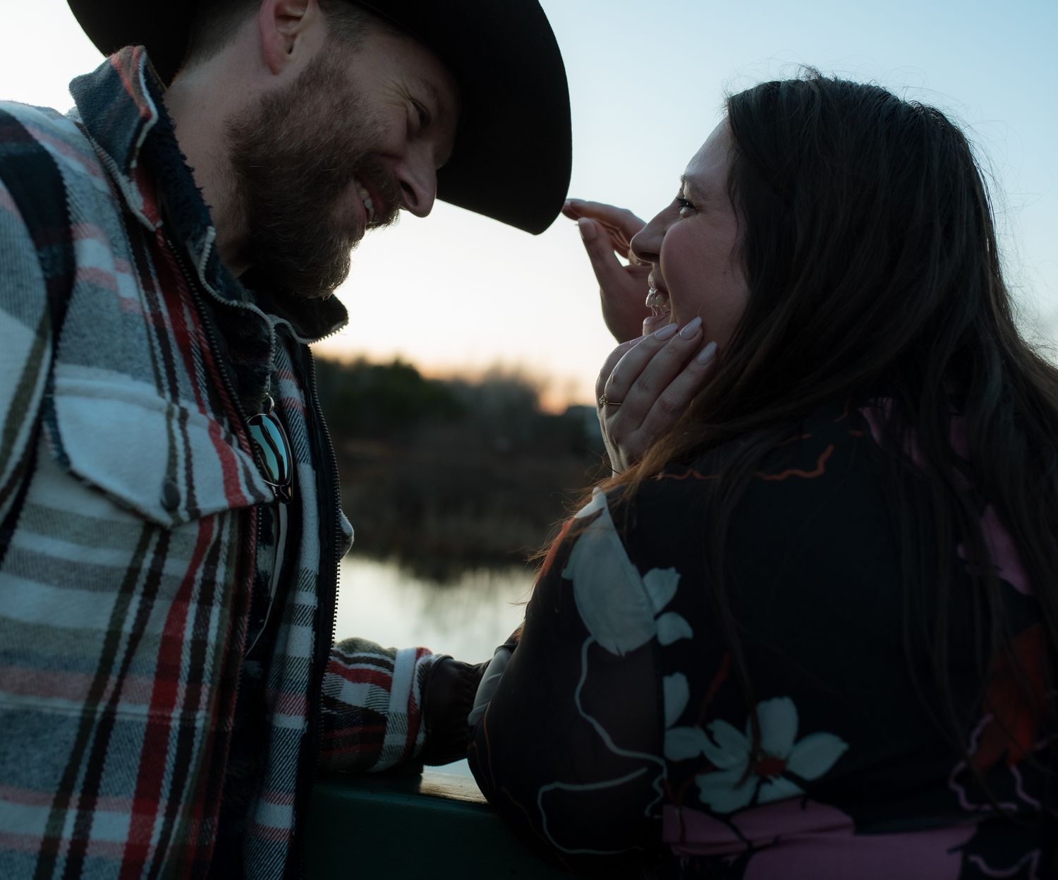 Two people in western wear share a tender moment against a waterfront backdrop at dusk.
