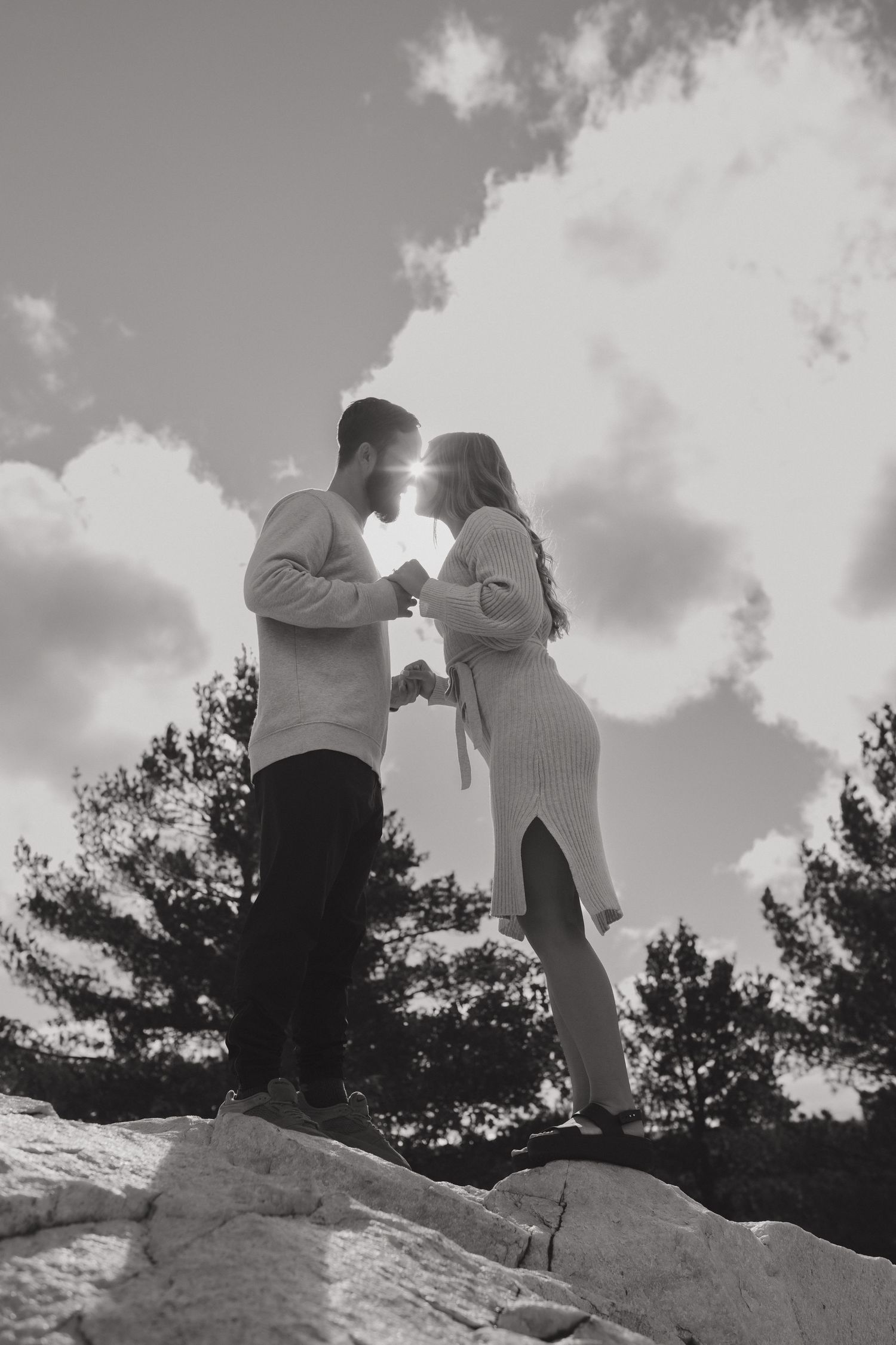 A romantic black and white silhouette of a couple standing on rocks with sunlit clouds in the background.