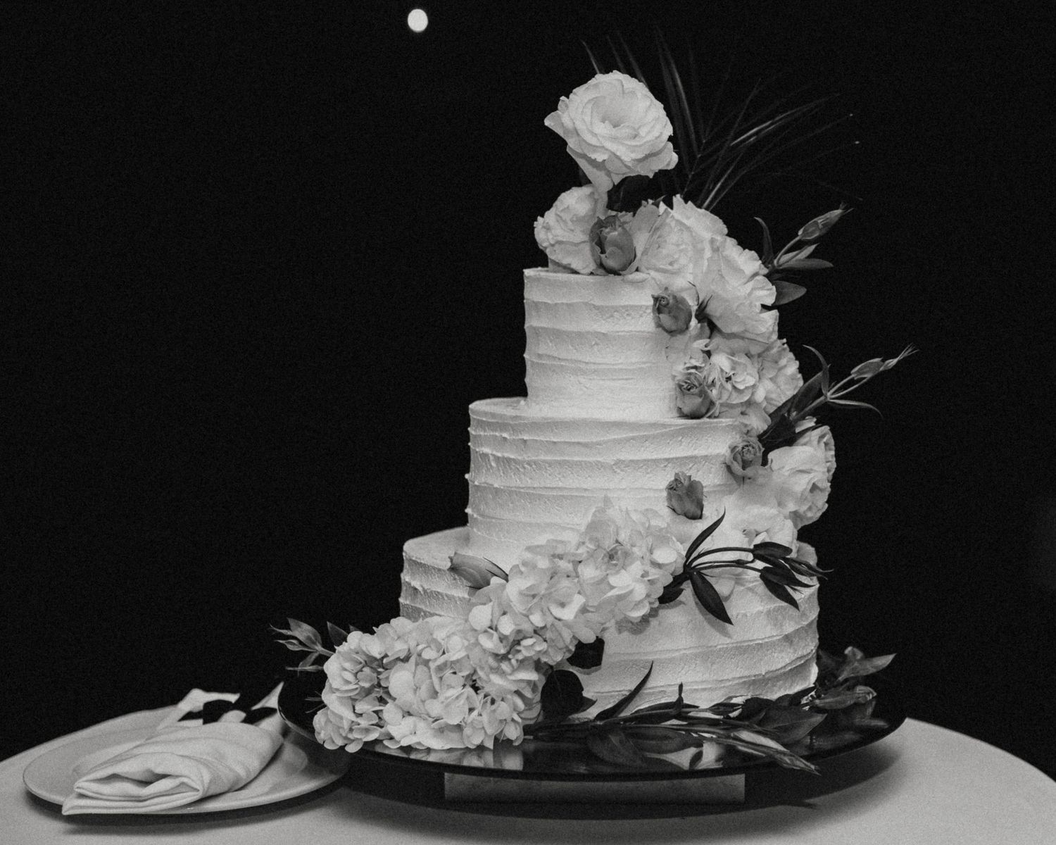 Elegant white wedding cake decorated with fresh roses and floral accents against dark background.