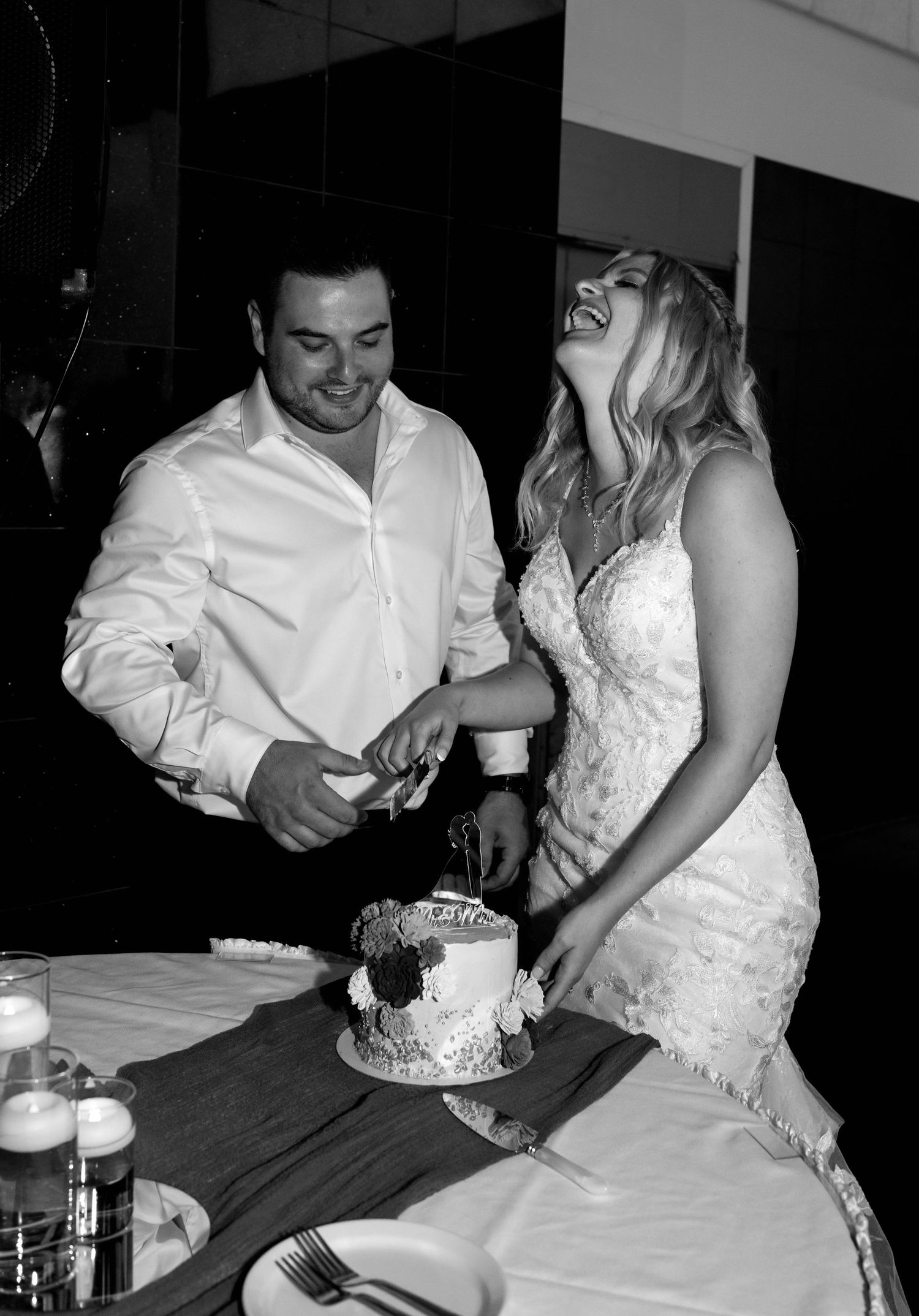 Black and white photo of a laughing bride in lace gown and groom cutting wedding cake at reception.