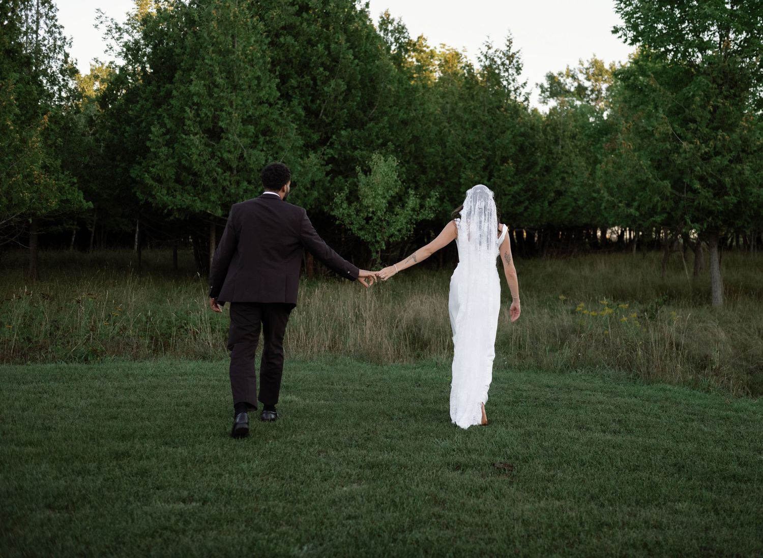 Bride and groom holding hands walking across green grass toward a tree-lined field at dusk.