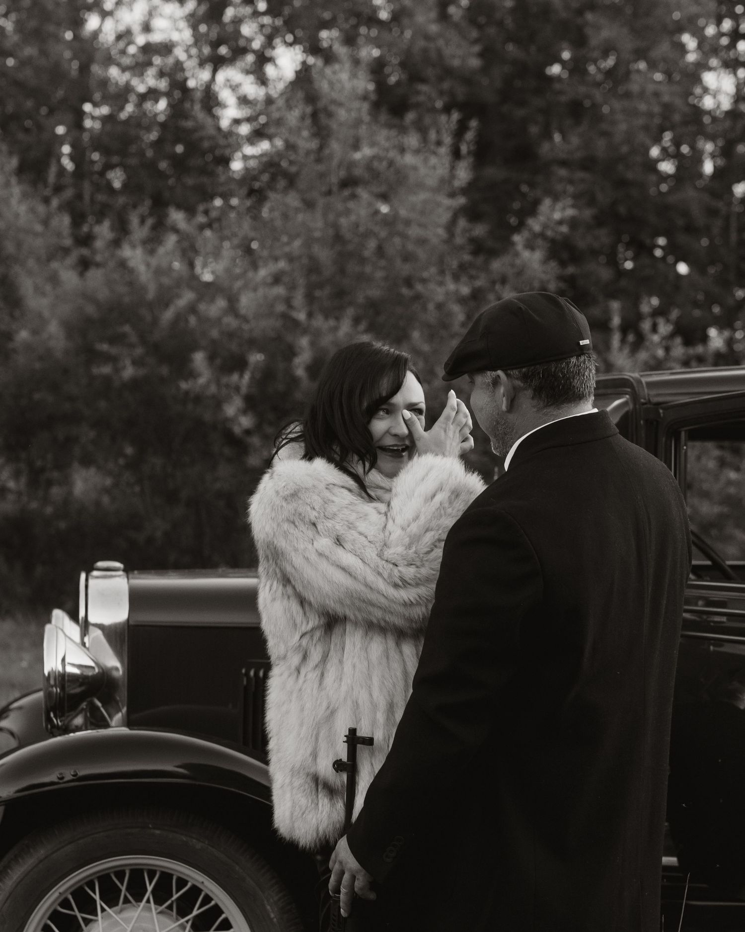 A black and white vintage photograph shows a couple in formal attire standing beside an antique automobile.