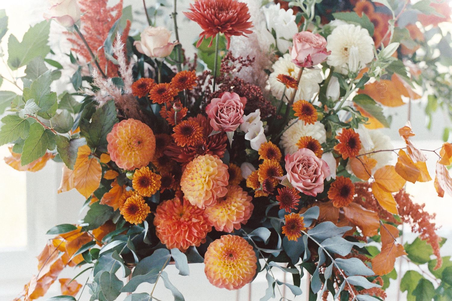 Colorful autumn floral arrangement with orange dahlias, pink roses, and greenery in rustic vases against white backdrop.