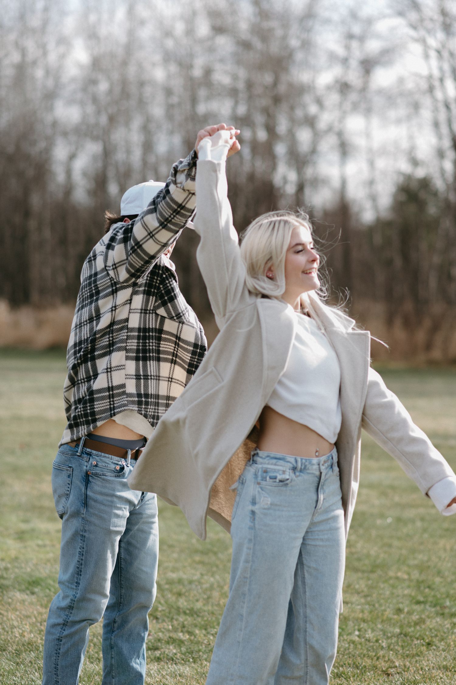 Couple dancing outdoors in jeans and casual jackets on a sunny day in a grassy field with bare trees.