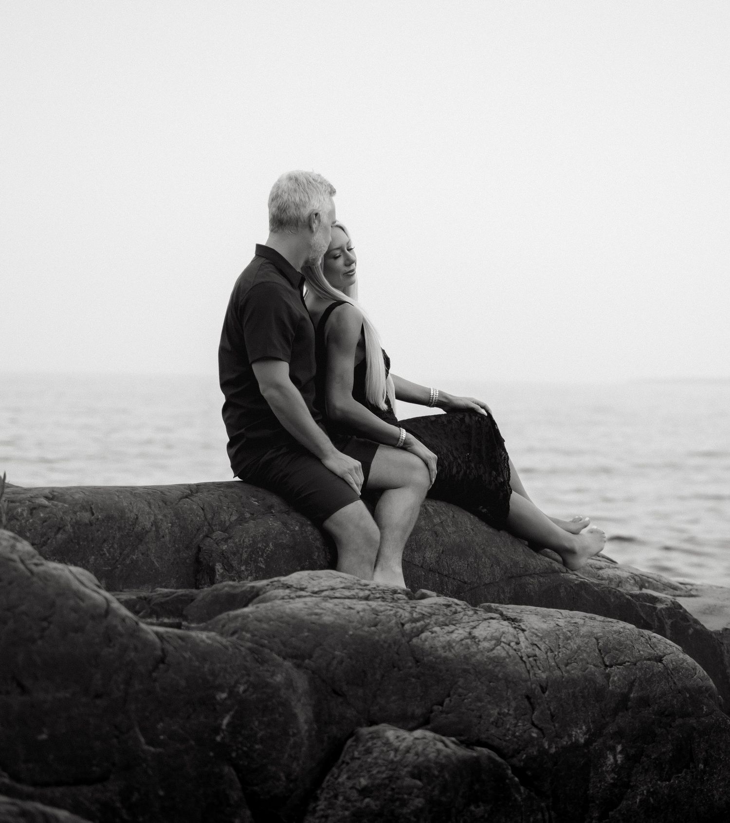 Black and white photo of a couple sitting on coastal rocks by the sea, sharing a romantic moment.