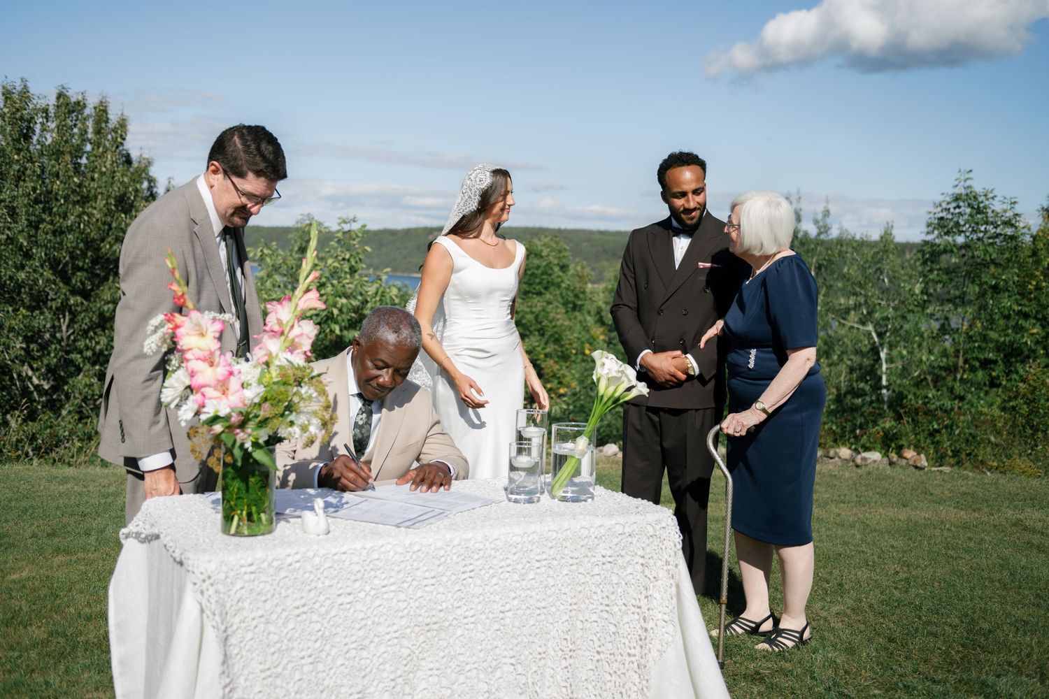 Outdoor wedding signing ceremony with bride, groom, officiant, and guests at a white table with floral arrangements.