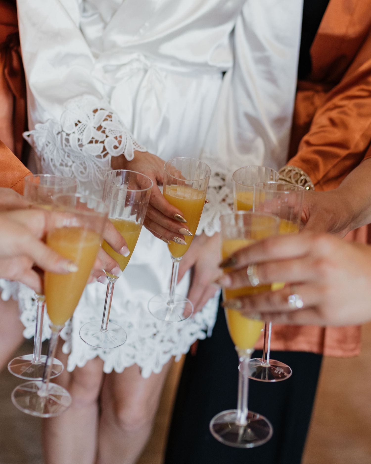 People toasting with mimosa cocktails in champagne flutes at a celebration with white lace dress details visible.