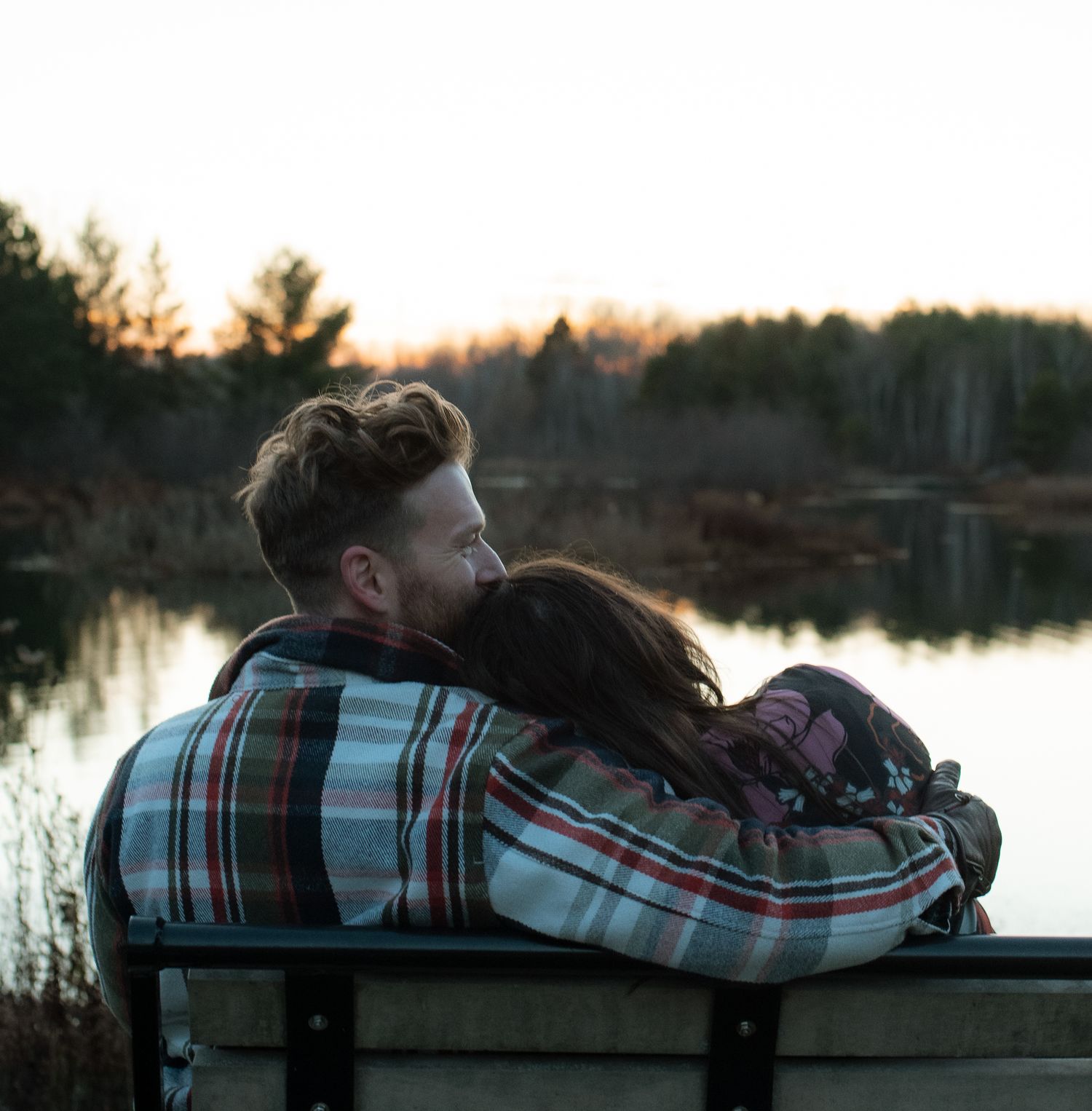 A couple in plaid clothing sits together on a bench overlooking a serene lake at sunset.