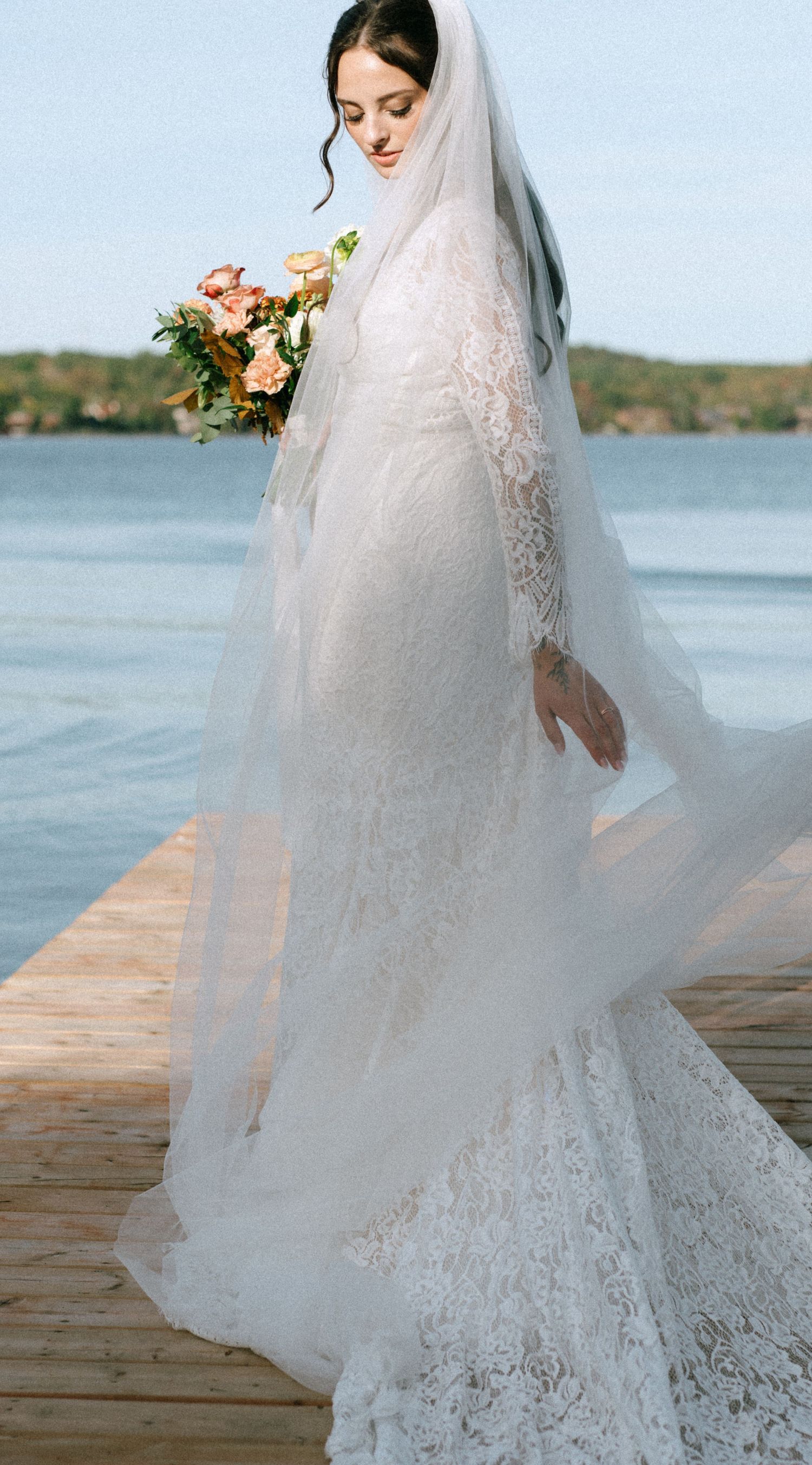 A bride in a lace wedding dress with veil stands on a wooden dock by the water.