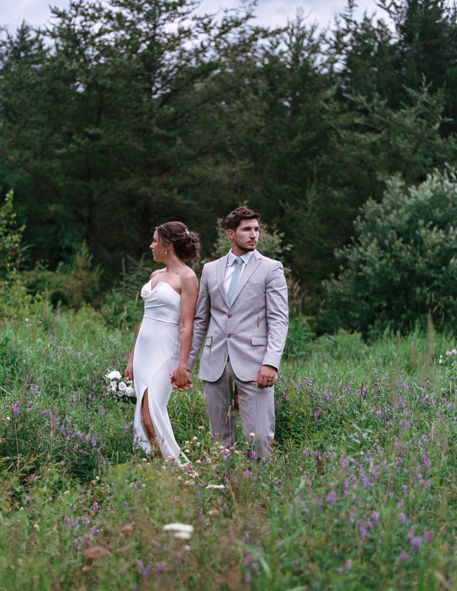 A bride in a white gown and groom in a gray suit hold hands in a lush wildflower meadow with evergreen trees behind them.