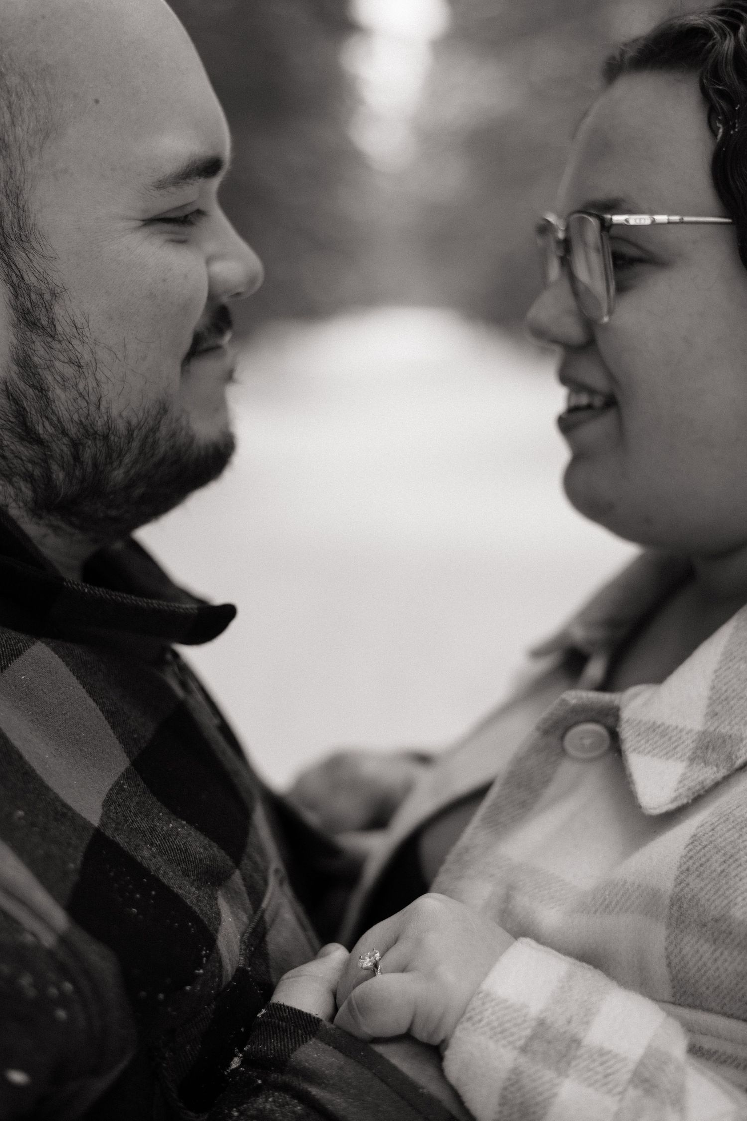 Black and white close-up of smiling couple facing each other, woman wearing glasses and engagement ring.