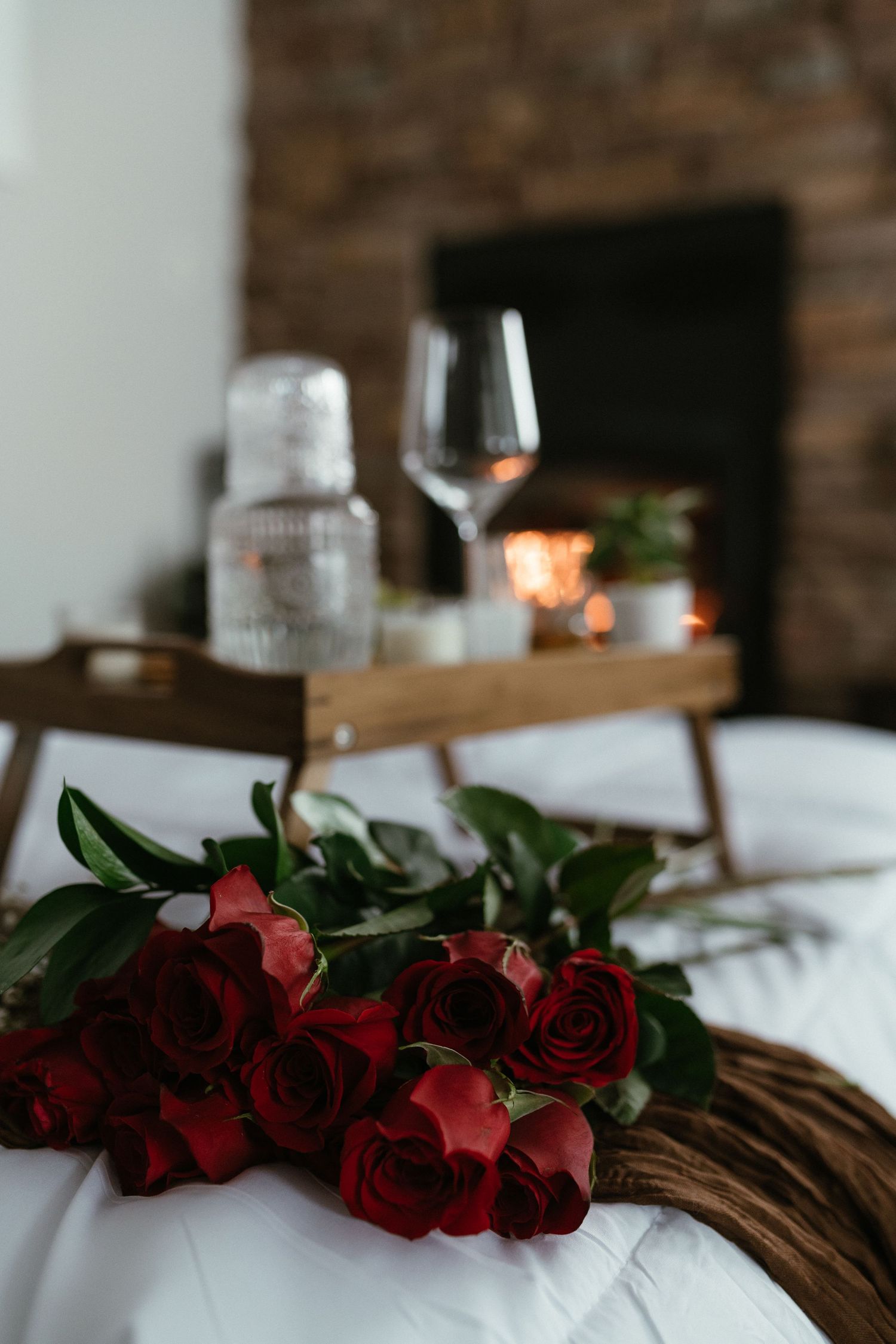 Bouquet of red roses on a white bed with a wooden tray, wine glass, and glowing candles in the background.