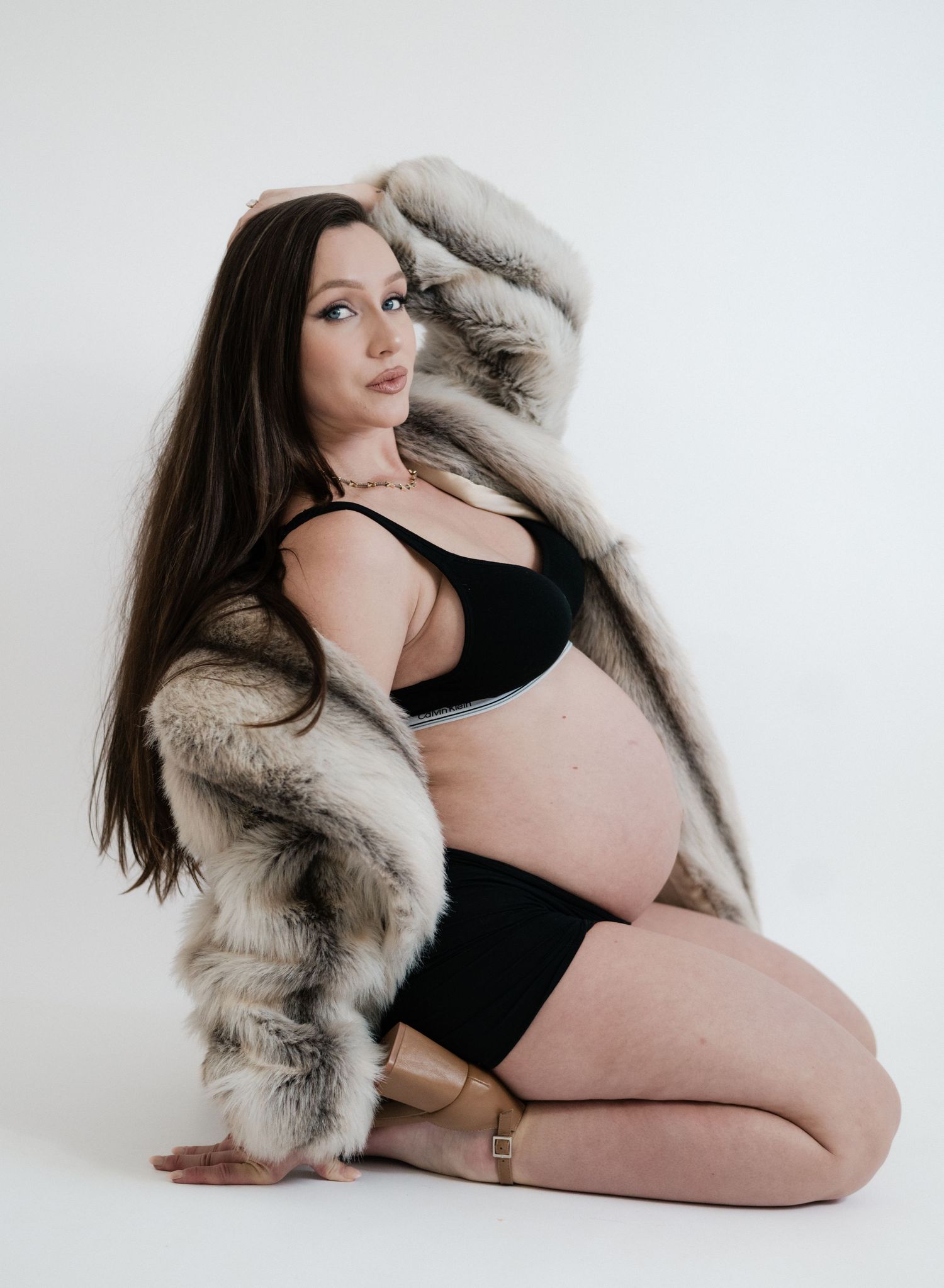 Pregnant woman in black bikini and fur coat posing on white background in elegant maternity photo shoot.