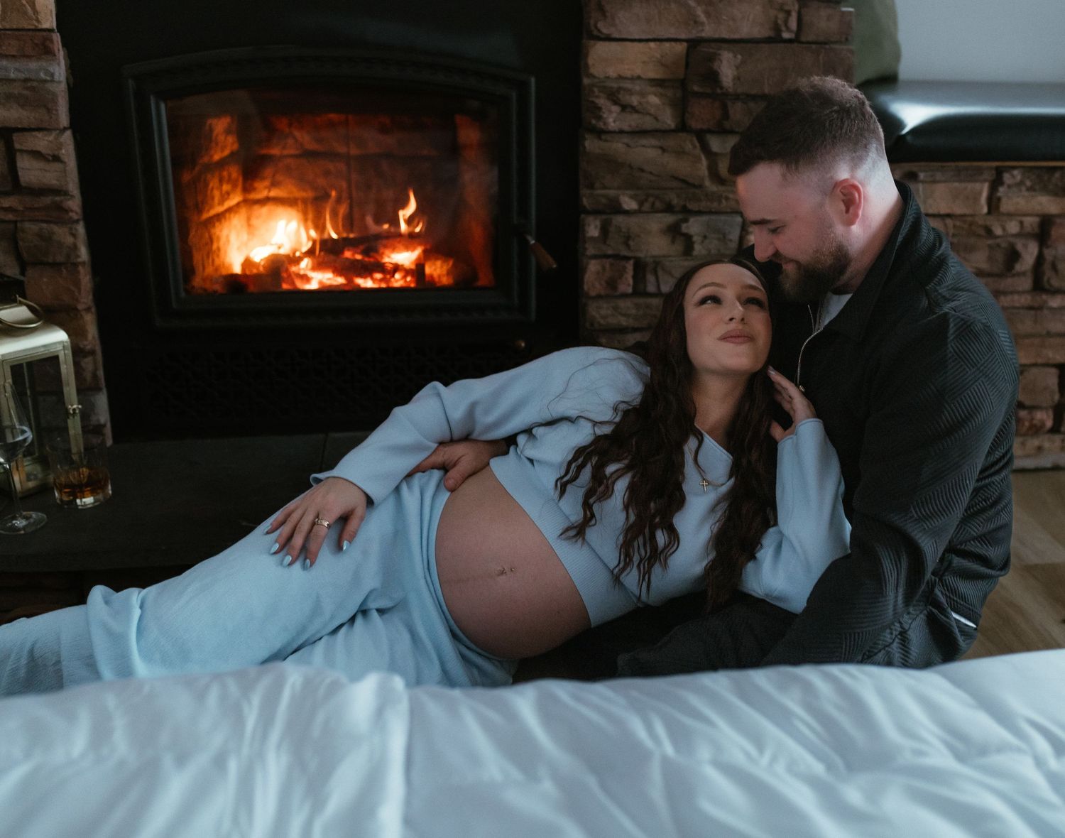Pregnant couple relaxing together on a bed by a warm glowing fireplace in a cozy stone-accented room.
