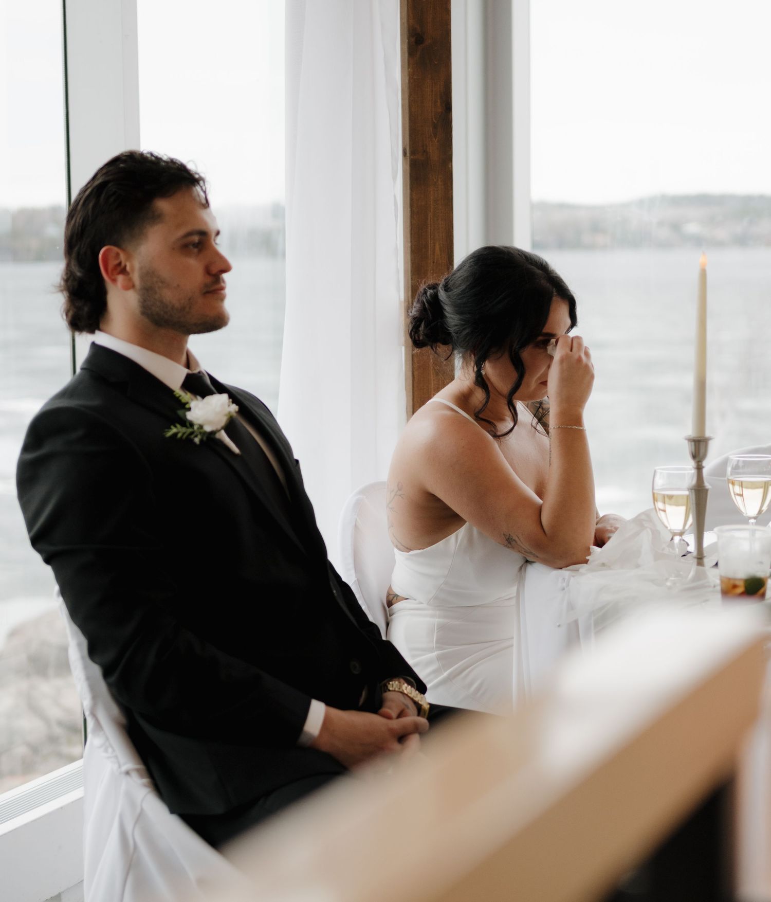 Emotional wedding reception moment with couple seated at head table by large windows overlooking water.
