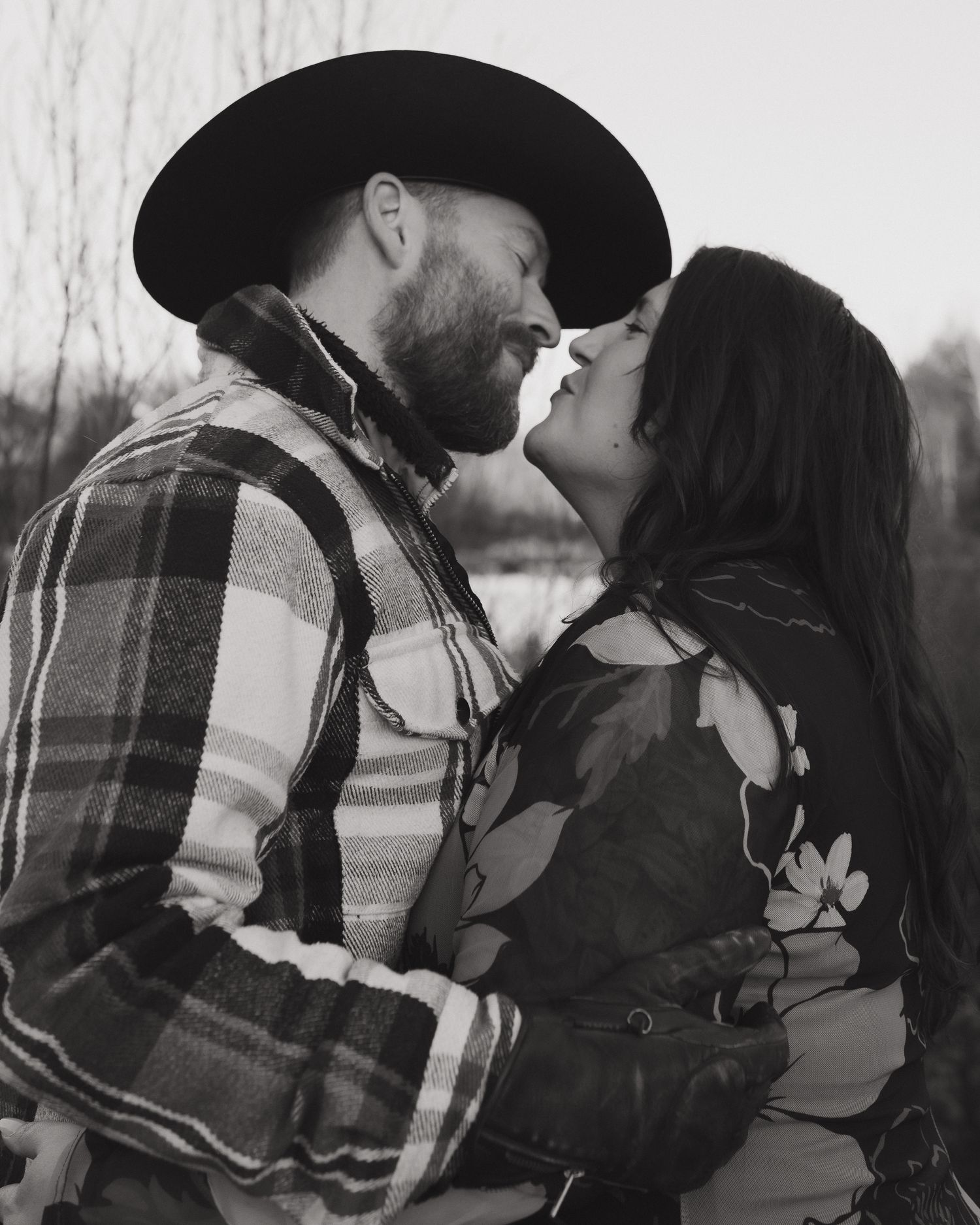 A couple in a plaid shirt and dark clothing share an intimate embrace in a black and white outdoor photoshoot.