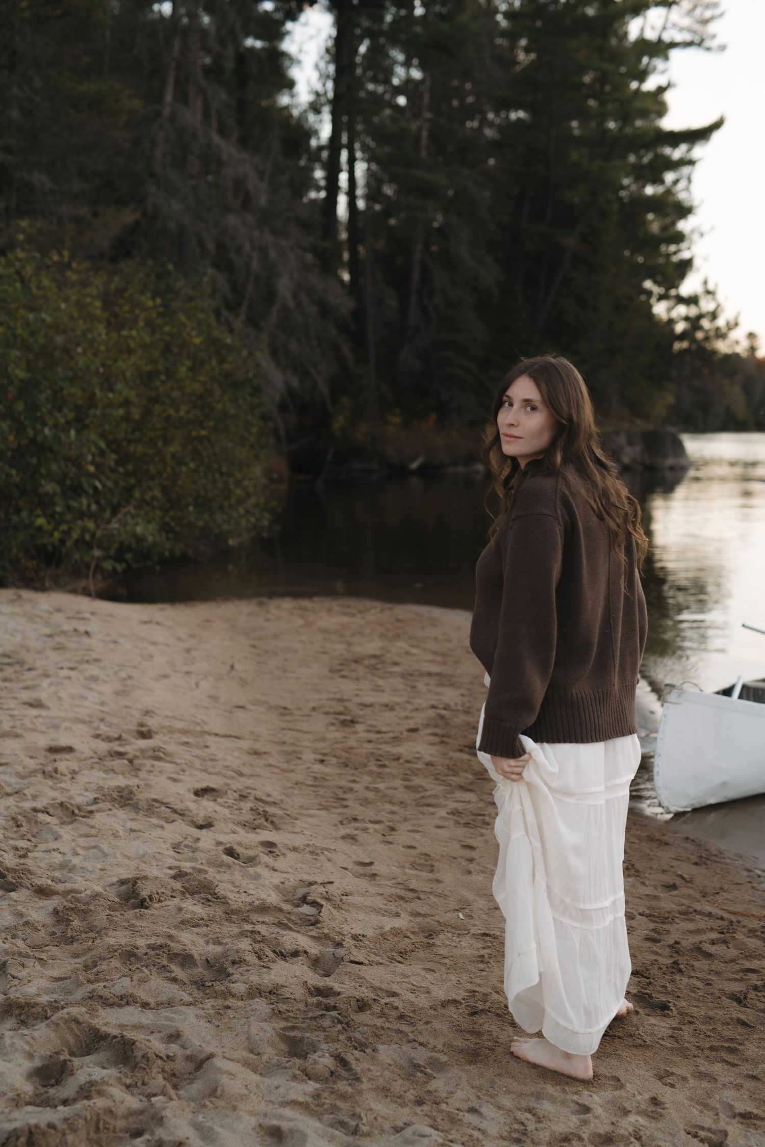 A couple stands on a sandy beach at sunset with evergreen trees and water in the background.